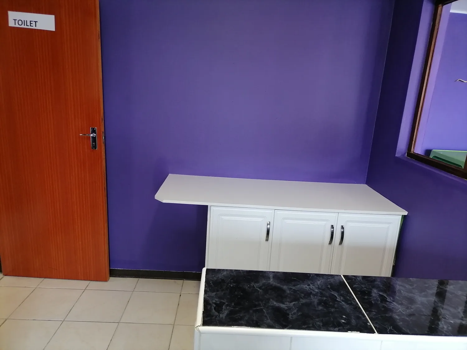 A purple-painted wash-up area with white cupboards, a long counter and a toilet door.