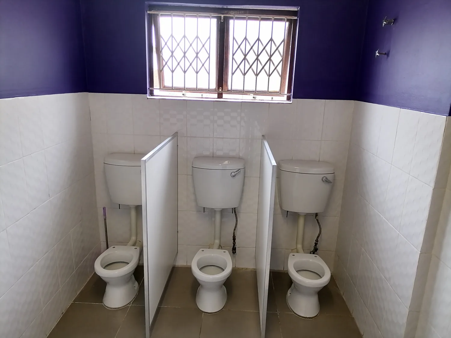 Three white toilets in the children's tiled washroom with a high window.